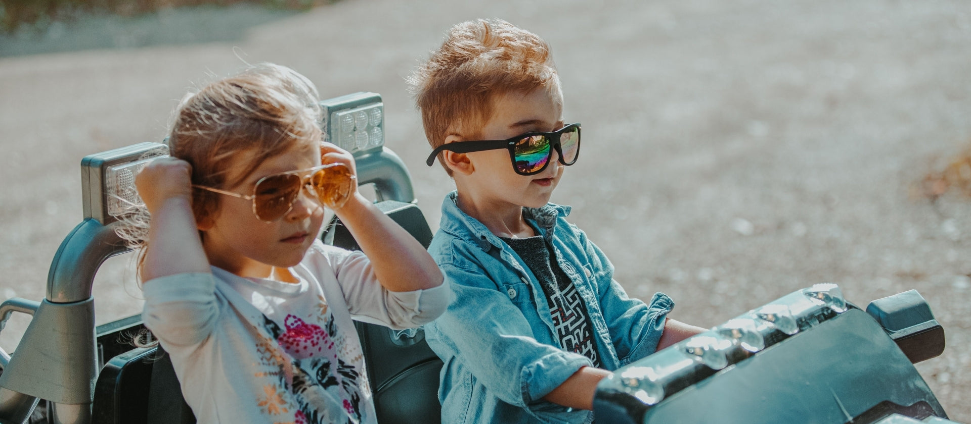 Two children wearing stylish sunglasses, one sitting in the passenger seat and one driving a small toy vehicle in an outdoor sandy setting.
