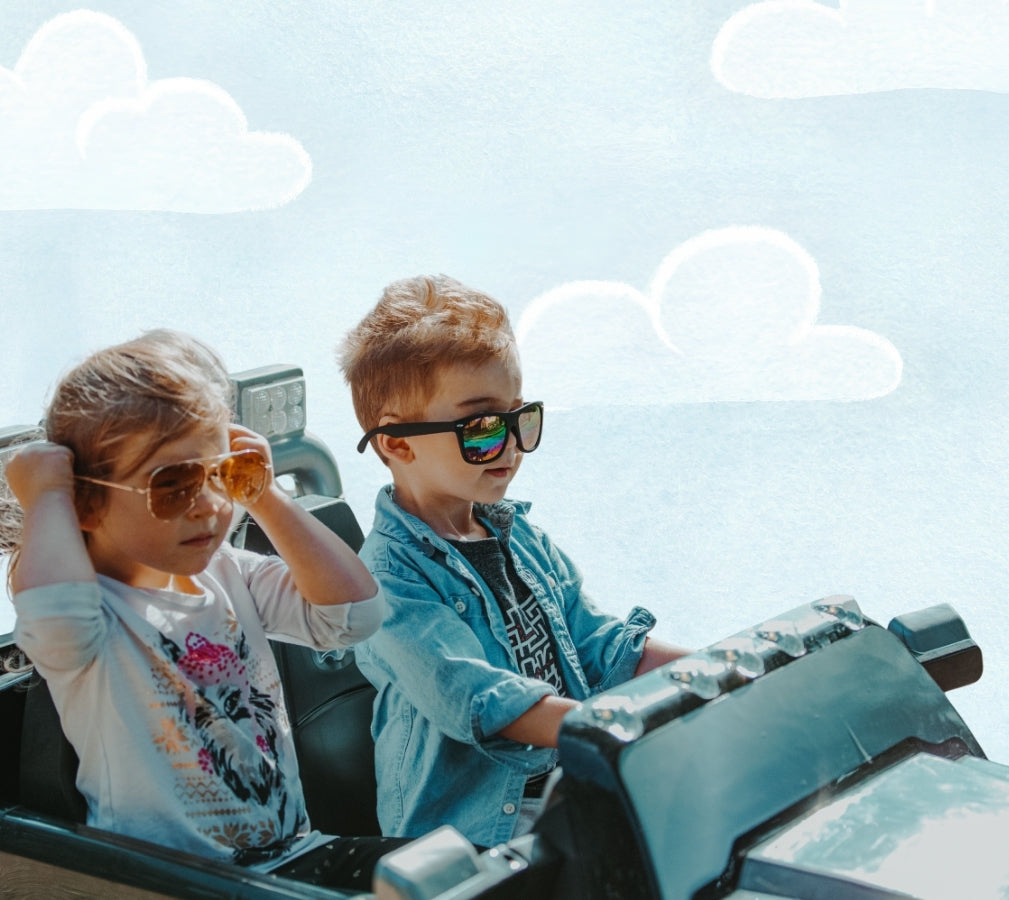 Two young kids wearing stylish sunglasses sitting in a ride vehicle, with a painted sky and clouds background.