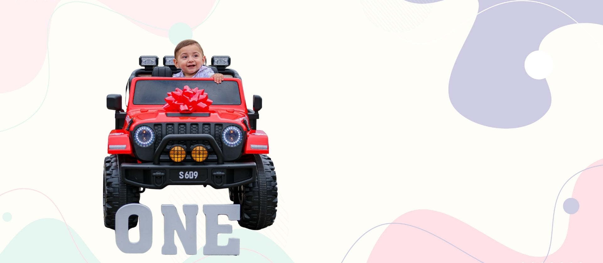 A child sitting in a red toy Jeep-style vehicle with a large red bow on the hood, set against a light background with abstract pastel shapes and the word 'ONE' at the bottom.